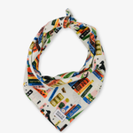 A cream-colored bandana or neck scarf featuring a colorful collage print of book spines, spiderwebs, crows, and labels, styled in a loop with a knot at the top on a light gray background.