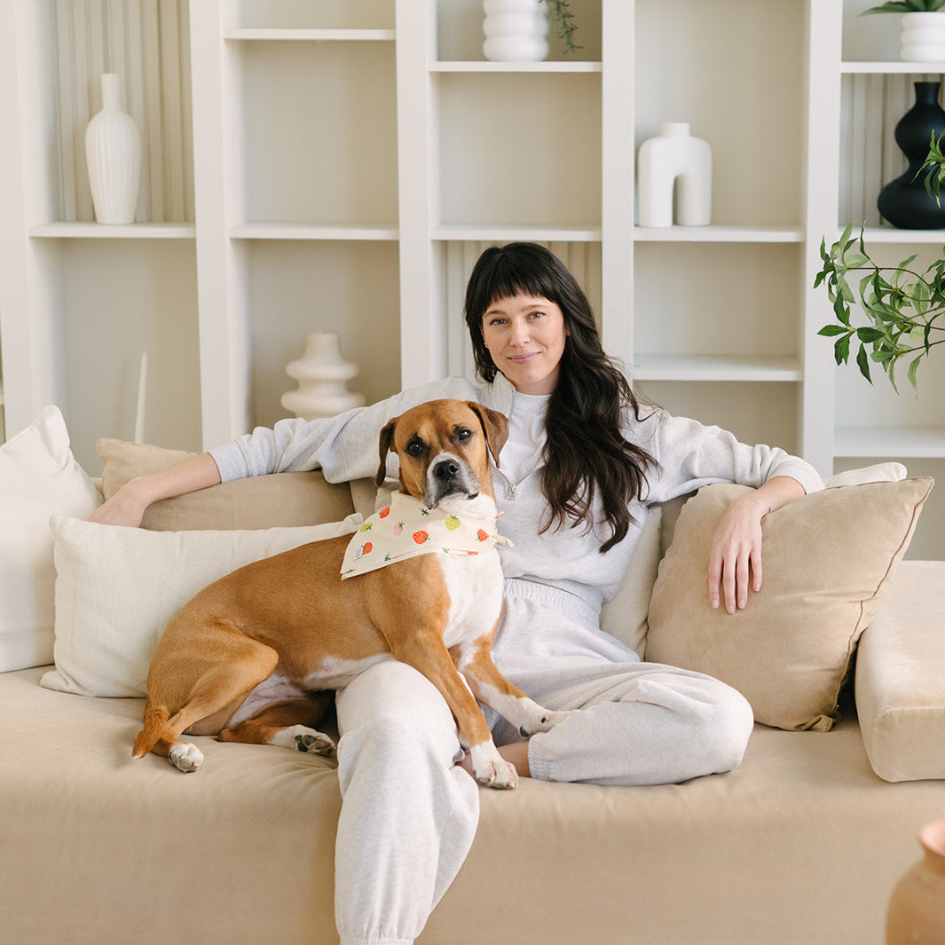 Woman sitting on a couch with a dog, surrounded by plants and shelves in a bright room.